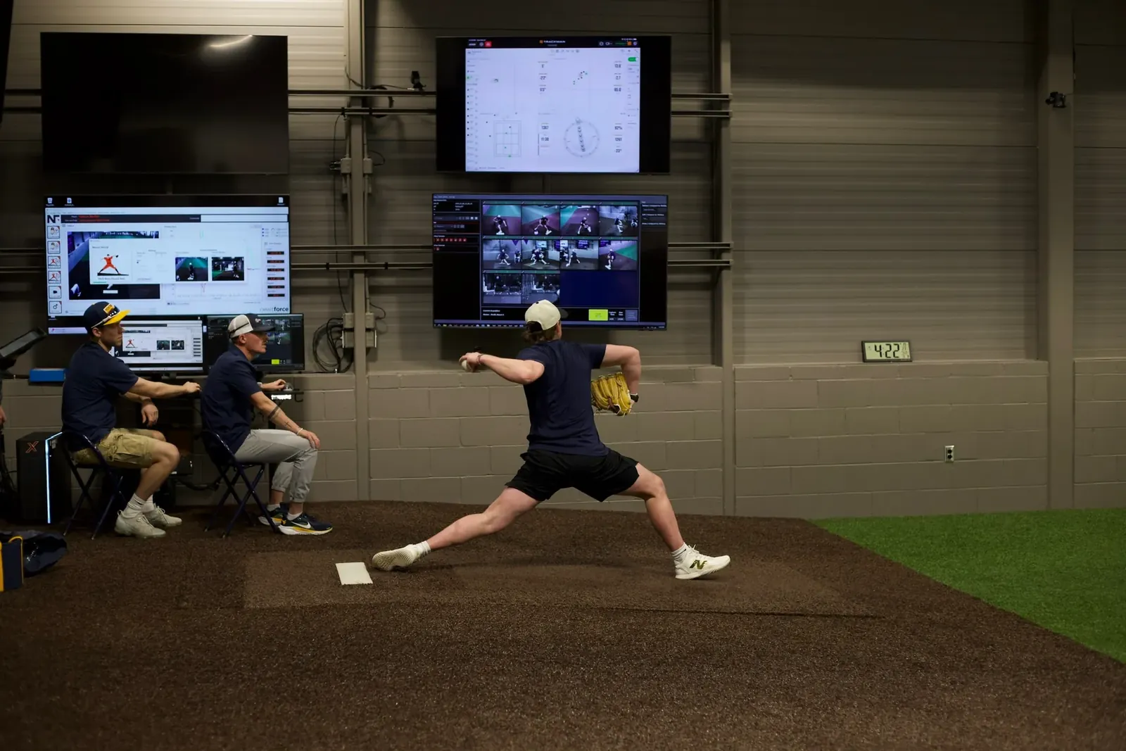 A pitcher mid-delivery on an indoor mound at BioPrecision's lab while two staff review pitch-analysis software on wall-mounted screens.